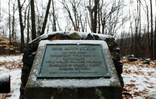 A stone monument with a plaque, surrounded by trees and snow, commemorating the Nine Men's Misery site.