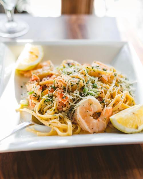 A plate of shrimp pasta topped with grated cheese and lemon wedges, served in a white dish on a wooden table.