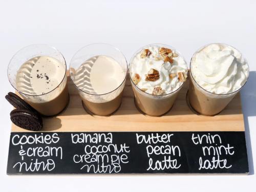 Four nitro lattes in clear cups on a wooden tray, labeled: Cookies & Cream, Banana Coconut Cream Pie, Butter Pecan, Thin Mint.