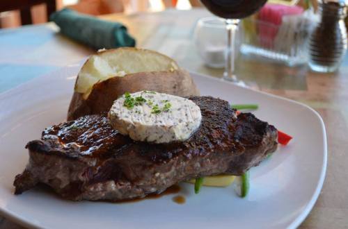 A grilled steak topped with herb butter, served with a baked potato and colorful vegetables on a white plate.