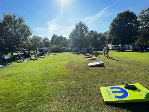 A sunny outdoor scene with people playing cornhole on a grassy field, surrounded by trees and parked vehicles.