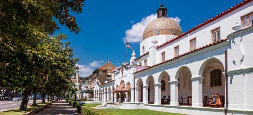 Historic building with a dome, arched windows, and a tree-lined street under a bright blue sky.