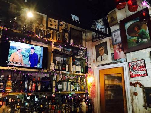 A cozy bar interior with a TV showing a classic film, surrounded by shelves of bottles and eclectic wall decor.