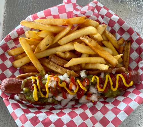 A hot dog topped with mustard, ketchup, onions, and relish, served with a side of golden French fries in a checkered basket.