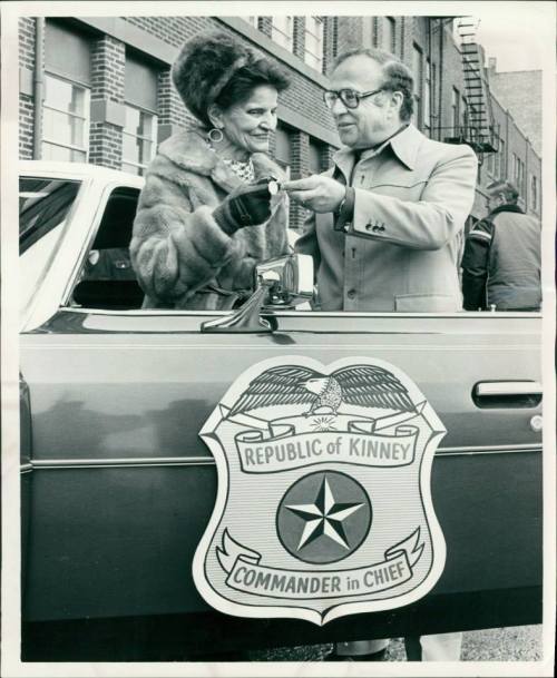 A man and woman exchange a key near a car with a "Republic of Kinney" emblem, smiling in a vintage black-and-white photo.