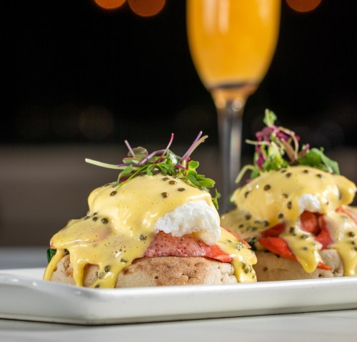 Two eggs Benedict on English muffins, topped with hollandaise sauce, strawberries, and microgreens, with a mimosa in the background.