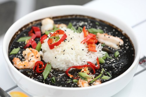 A bowl of black seafood soup with shrimp, white rice, red chili slices, and green onions, garnished with sesame seeds.