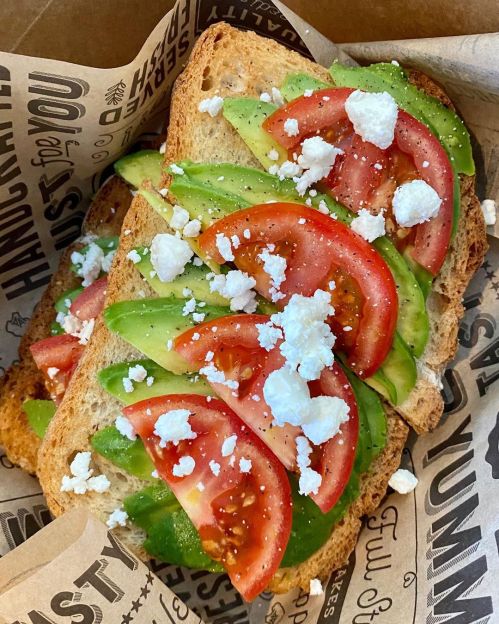 A close-up of a sandwich with avocado, tomato slices, and crumbled feta cheese on whole grain bread.