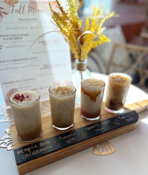 A wooden flight board holds five different drinks, each in a small glass, with a decorative vase and menu in the background.