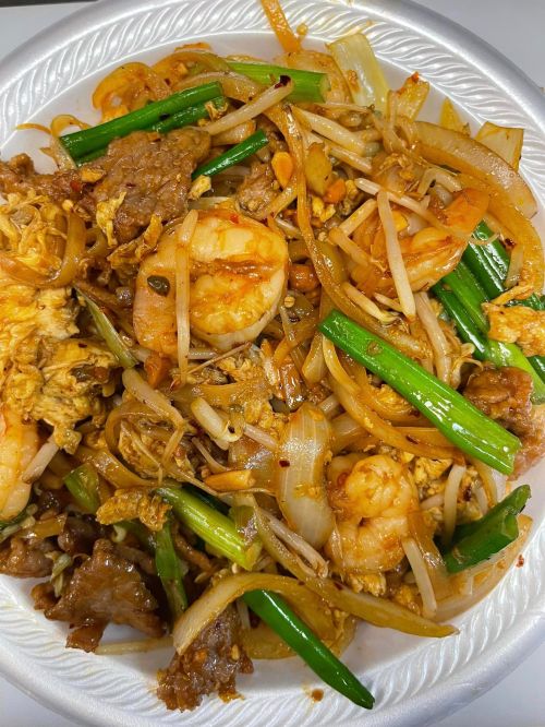 A plate of stir-fried noodles with shrimp, beef, onions, green onions, and bean sprouts, garnished with spices.