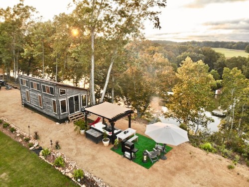 A tiny house by a river, surrounded by trees, with a patio area featuring seating and an umbrella.