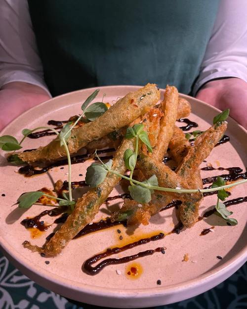 A plate of crispy fried green beans drizzled with sauce and garnished with microgreens, held by a person.