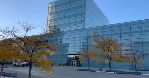 Modern glass building with the word "FISGE" visible, surrounded by trees with yellow leaves. Clear blue sky in the background.