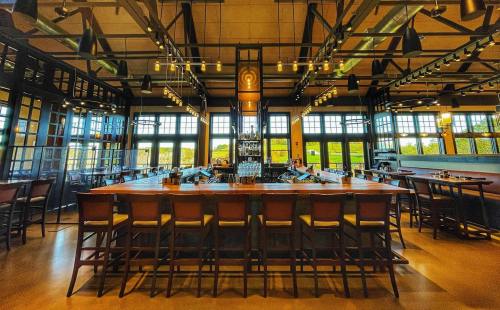 A spacious restaurant interior featuring a large wooden bar, high stools, and large windows with natural light.