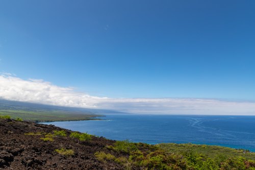 A scenic view of a coastline with blue ocean, green land, and a clear sky, framed by volcanic rock.