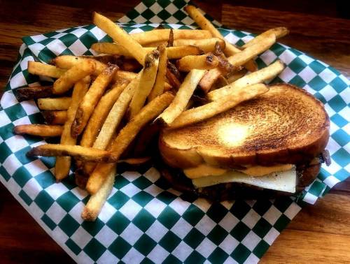 A toasted sandwich with cheese and pickles, served with a side of golden French fries on a checkered paper.