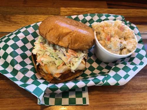 A sandwich with coleslaw on a bun, served with a side of creamy pasta salad in a cup, on a checkered paper.