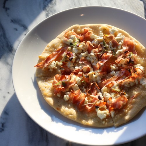 A flatbread topped with cheese, ham, and herbs, served on a white plate against a marble background.