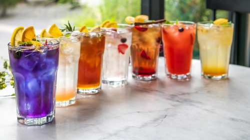 A row of colorful cocktails in tall glasses, garnished with fruit and herbs, on a marble table.