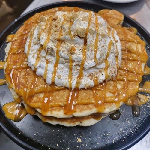 A stack of pancakes topped with whipped cream, caramel drizzle, and crushed graham crackers on a black plate.