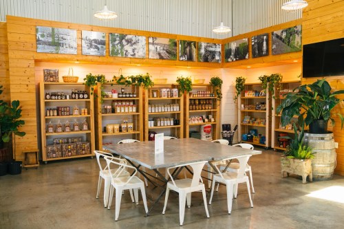 A cozy shop interior with wooden shelves filled with products, a metal table, and white chairs surrounded by plants.