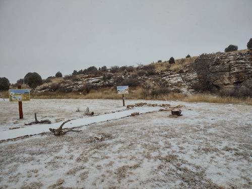 A snowy landscape with signs and rocky terrain, surrounded by sparse vegetation and a hill in the background.