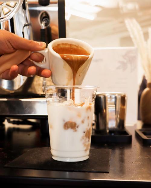 A hand pours espresso from a cup into a glass of iced milk, creating a layered coffee drink.