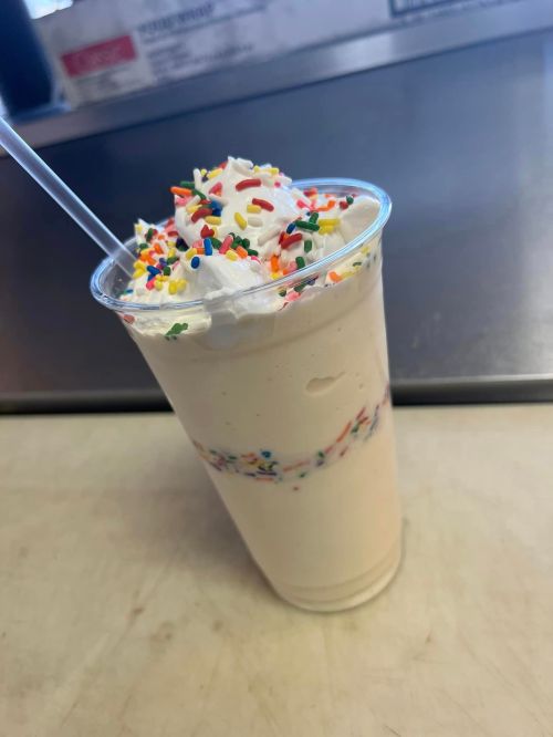 A creamy milkshake topped with whipped cream and colorful sprinkles in a clear cup with a straw.