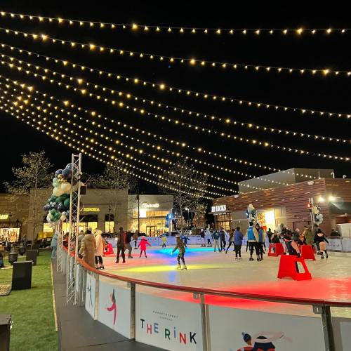 A lively outdoor ice skating rink at night, adorned with string lights and surrounded by shops and festive decorations.