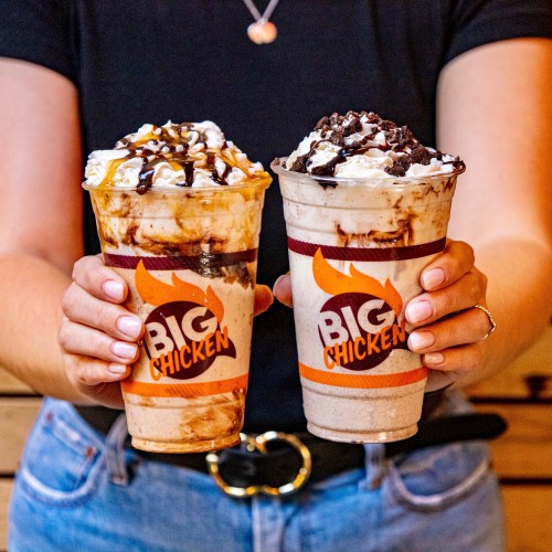 A person holds two large milkshakes from Big Chicken, one topped with caramel and the other with chocolate and cookies.