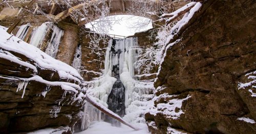 A frozen waterfall surrounded by snow-covered rocks and trees, with a stone bridge above.