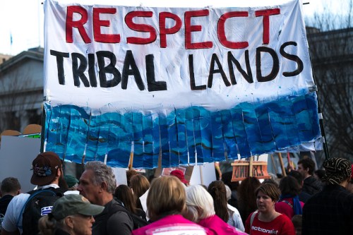 A crowd gathers under a large banner reading "Respect Tribal Lands," advocating for Indigenous rights.