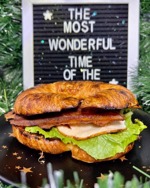 A croissant sandwich with lettuce, turkey, and bacon, beside a sign that reads "The Most Wonderful Time of the."