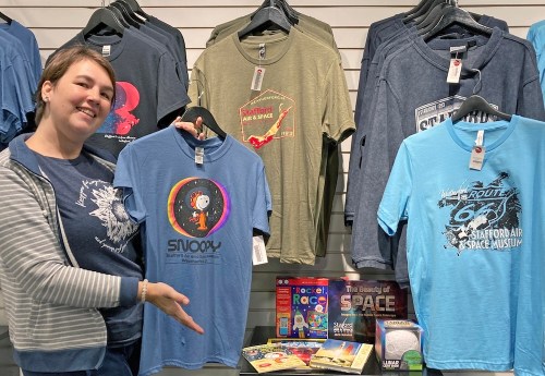 A person points to a blue Snoopy t-shirt among various other shirts displayed on a wall.