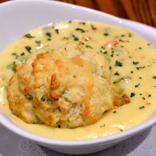A bowl of creamy soup topped with a fluffy biscuit, garnished with herbs.