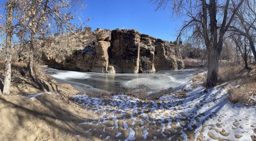A serene winter landscape featuring a frozen lake surrounded by rocky cliffs and bare trees under a clear blue sky.