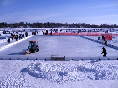 A busy outdoor ice rink with skaters, snow, and a zamboni in a winter landscape under a clear blue sky.