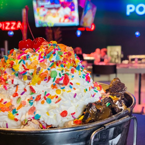 A large dessert bowl filled with ice cream, whipped cream, sprinkles, and cherries, set in a vibrant restaurant.