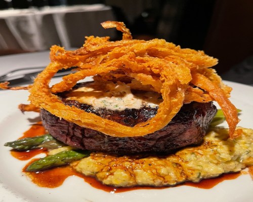 A gourmet dish featuring a grilled steak topped with crispy onions, served on a bed of asparagus and creamy sauce.