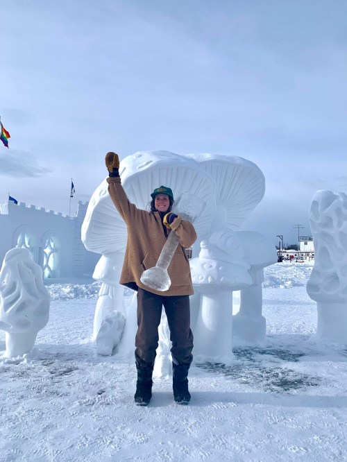 A person in winter clothing stands proudly next to a large snow sculpture, raising a snow tool in celebration.