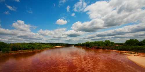 A serene river flows through lush greenery under a bright blue sky with fluffy clouds.