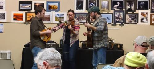 Three musicians perform with a mandolin, violin, and guitar in front of a wall of framed photographs. Audience members watch.