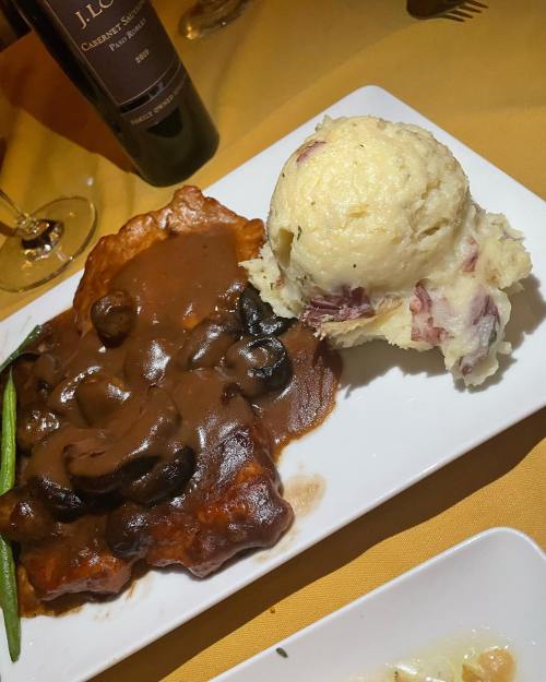 A plate featuring chicken with mushroom sauce, mashed potatoes, and a side of green beans, with a wine bottle in the background.