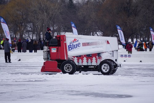 A Zamboni cleans ice at an outdoor rink, with spectators and banners in the background.