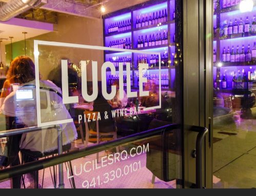 A view of Lucile Pizza & Wine Bar through a glass door, featuring a vibrant purple-lit wine display in the background.