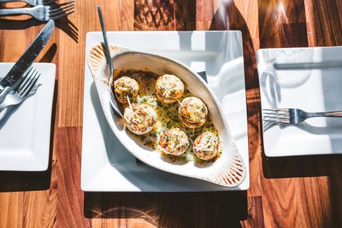 A dish of baked seafood balls topped with cheese, served on a white plate, with cutlery and empty plates nearby.
