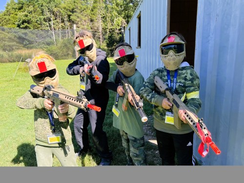 Four children in masks and tactical gear pose with toy guns in an outdoor setting, ready for a game.