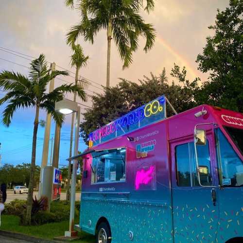 A colorful food truck parked under a rainbow, surrounded by palm trees and a vibrant sky.