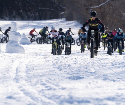 A group of cyclists riding through snowy terrain, with a snowman in the foreground.