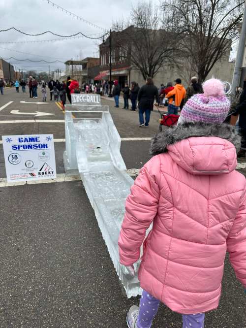 A child in a pink coat approaches an ice slide at a winter event, with a crowd and sponsor sign in the background.